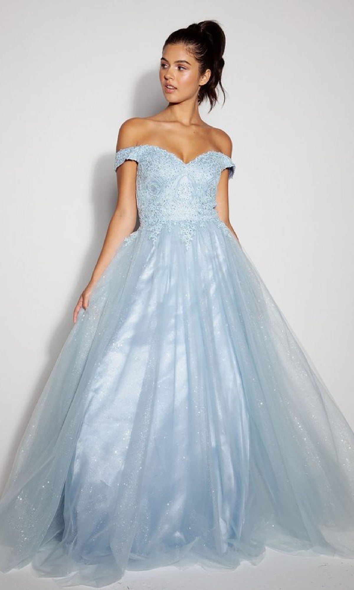 A woman wears the Off-Shoulder Glitter Prom Ball Gown: Eureka 9899, featuring a light blue off-the-shoulder design with a fitted bodice, lace details, and a full tulle skirt.
